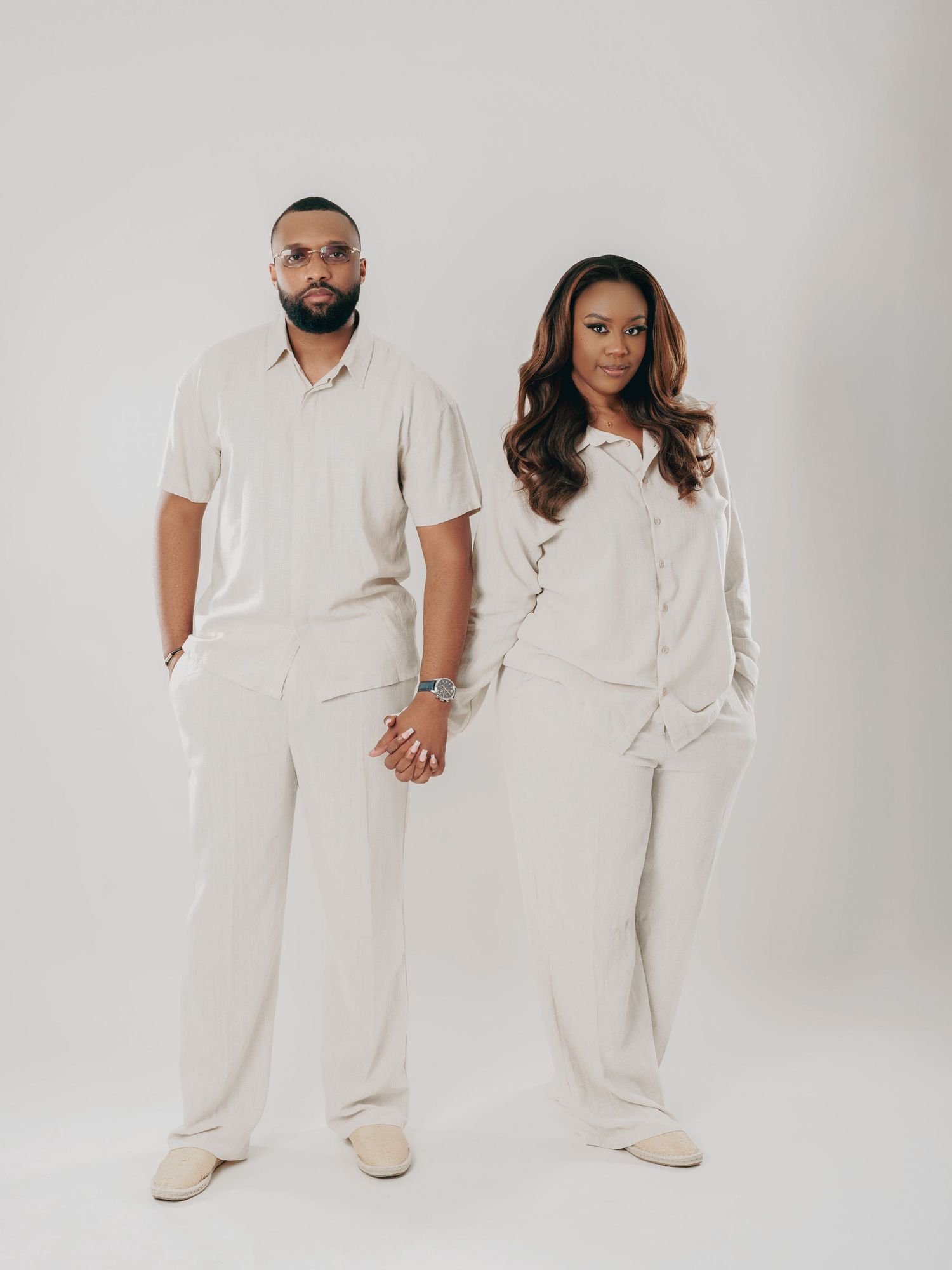 Couple holds hands wearing coordinated beige linen outfits in a minimalist studio setting.