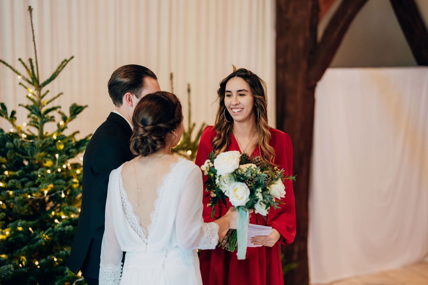 Bridesmaid in red dress holding white bouquet smiling at bride during winter wedding ceremony.