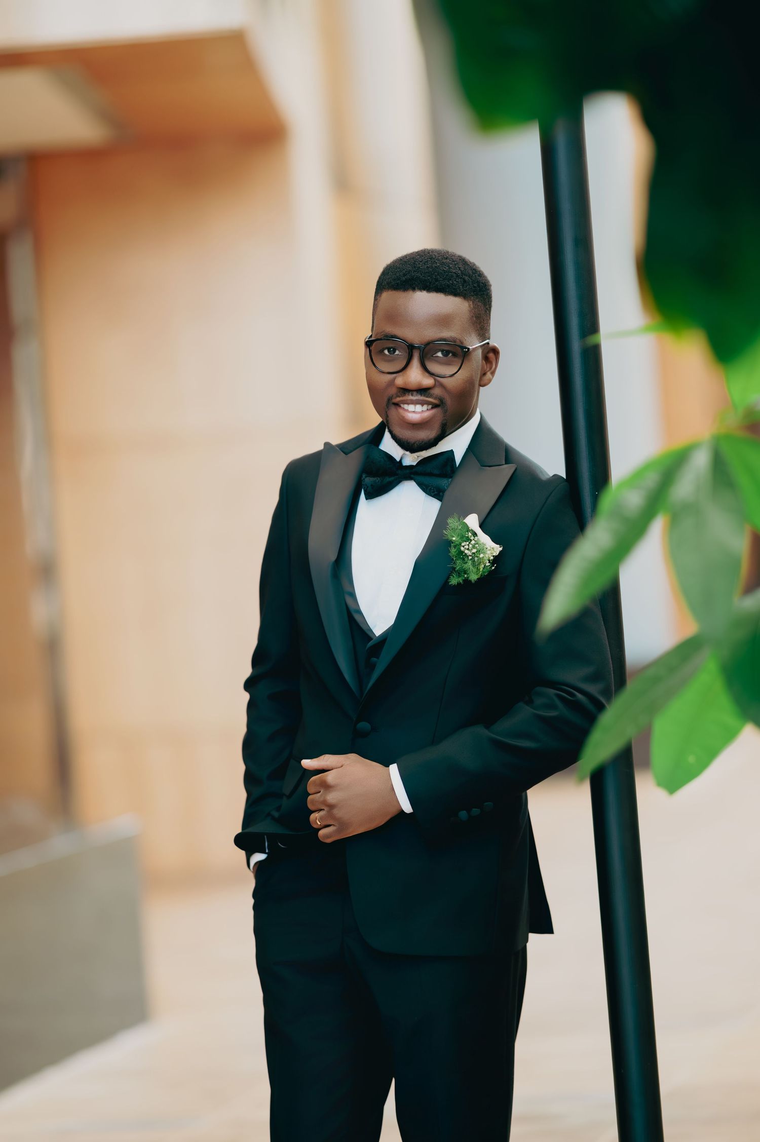 Smiling groom in black tuxedo with boutonniere posing outdoors near green foliage.