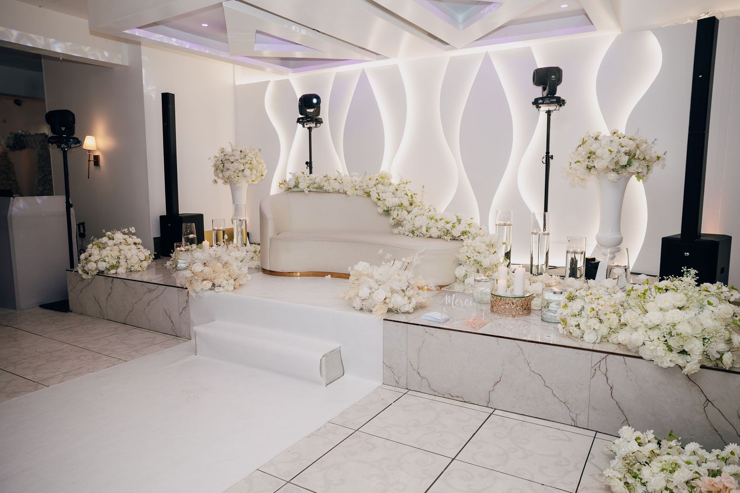 All-white wedding reception venue with marble stage, white floral arrangements, and modern wave-pattern wall backdrop.
