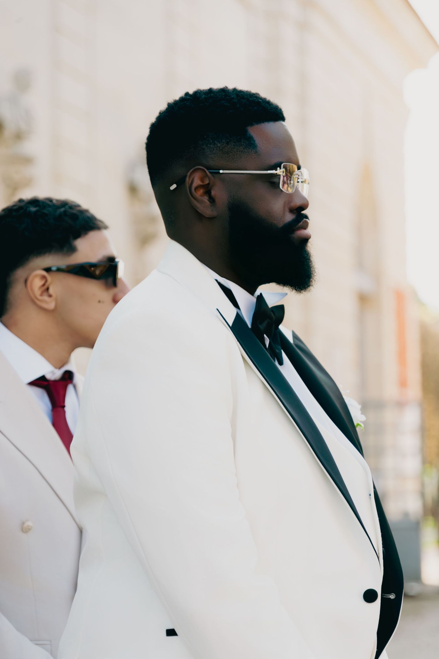 Dapper groom in white tuxedo with black bow tie and stylish glasses at wedding venue.