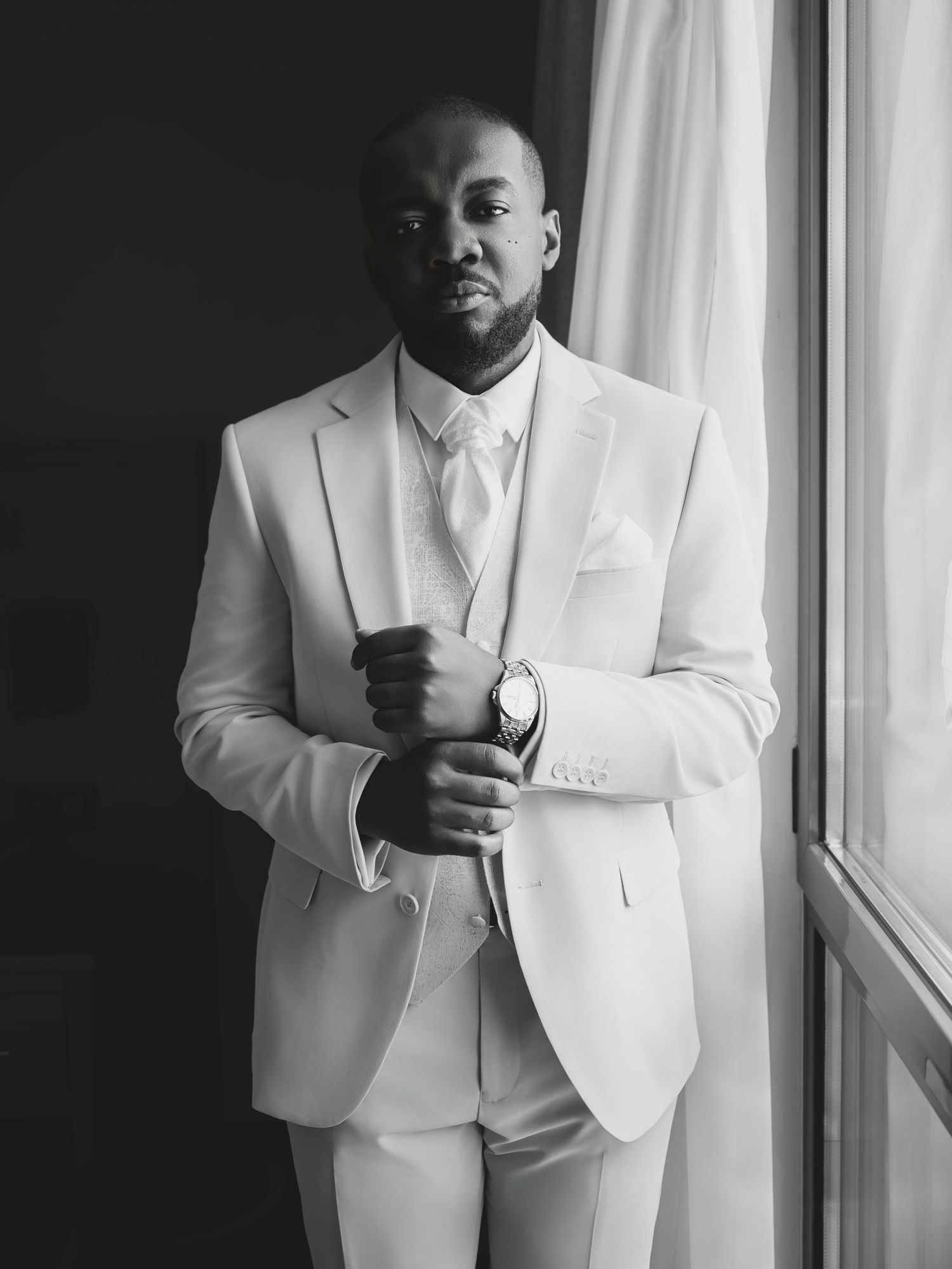 Elegant man in white suit adjusting cufflinks by window in black and white portrait photo.