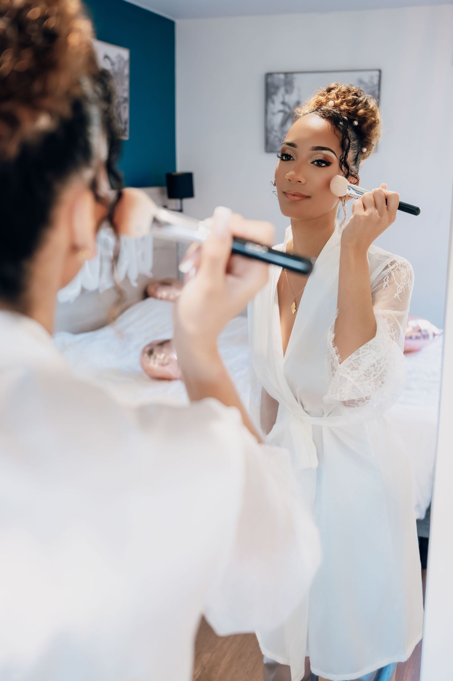 Bride in white lace robe applying makeup with brush while getting ready for wedding day.