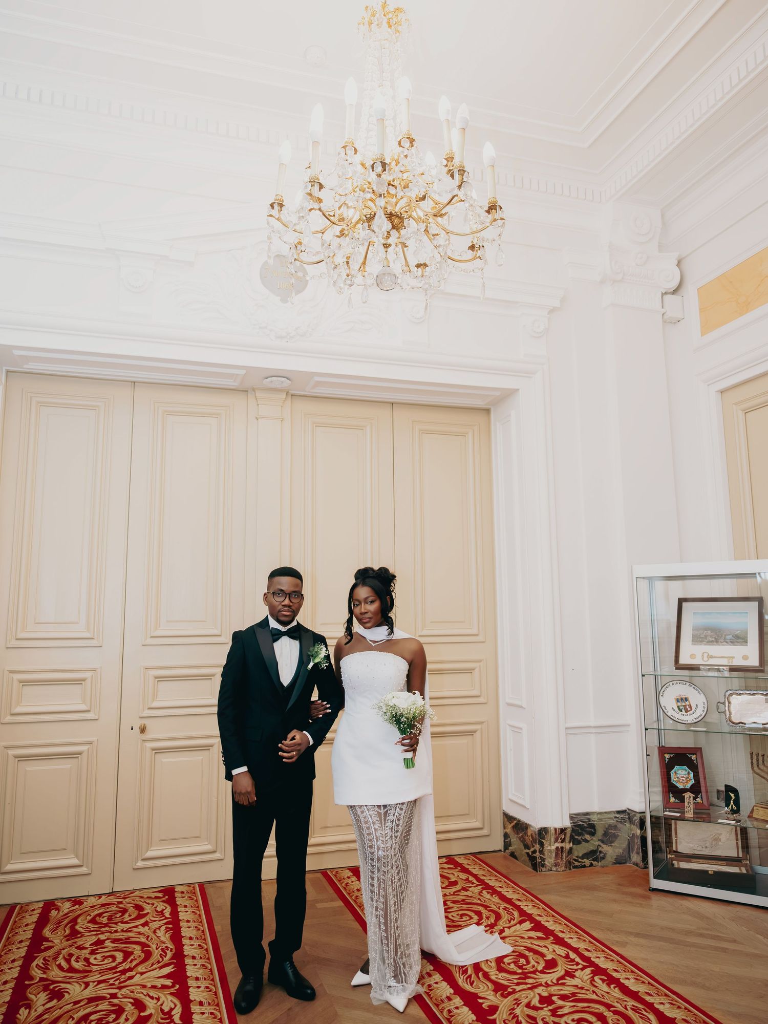 Newlywed couple poses under gold chandelier in grand white French chateau wedding venue hall.