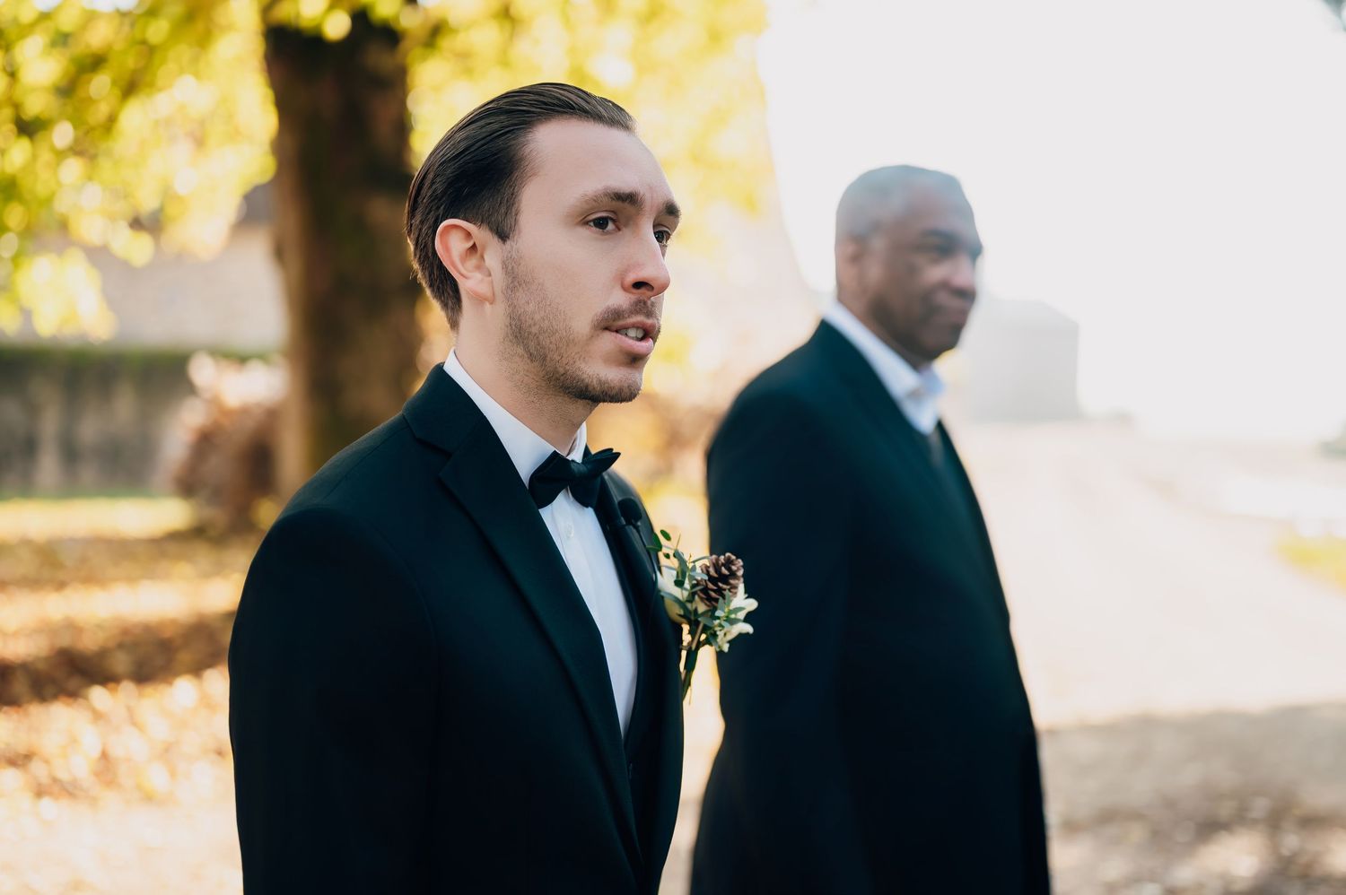Handsome groom in black tuxedo with bow tie and floral boutonniere standing outdoors during autumn wedding ceremony.