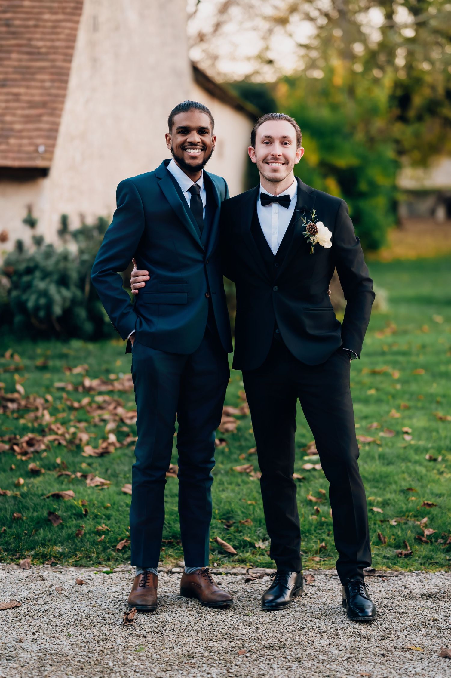 Two smiling groomsmen in dark suits pose together outdoors, one wearing black tuxedo with boutonniere.