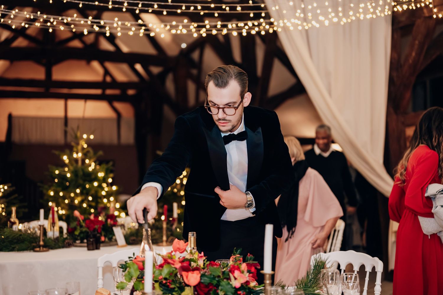 Elegantly dressed man in tuxedo lighting candles at a Christmas wedding reception table with festive red floral arrangements.