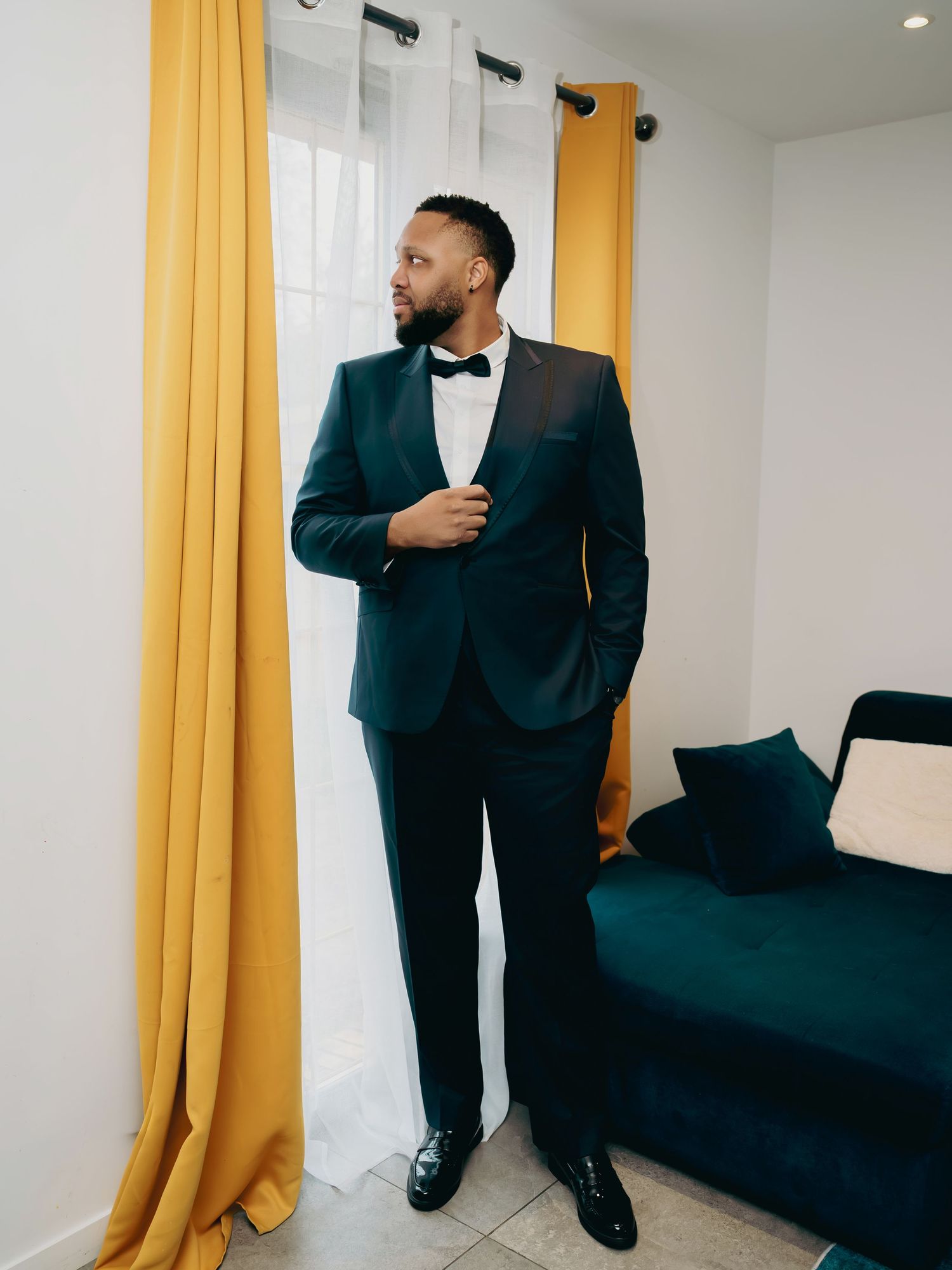 Handsome Black groom in dark teal tuxedo with bow tie posing by yellow curtains before wedding.