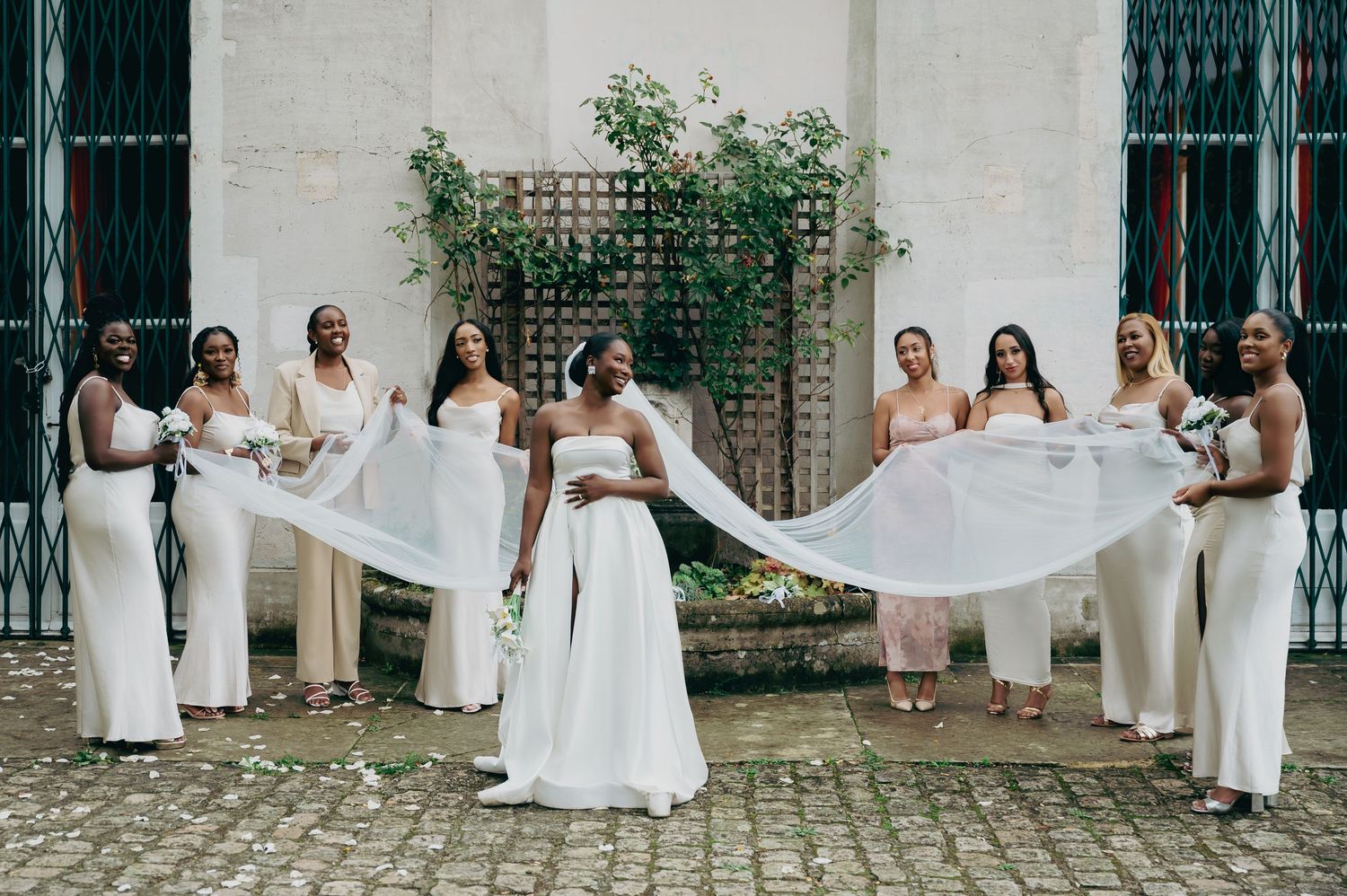 Bride in white gown with flowing veil extended by bridesmaids in ivory dresses at wedding venue.