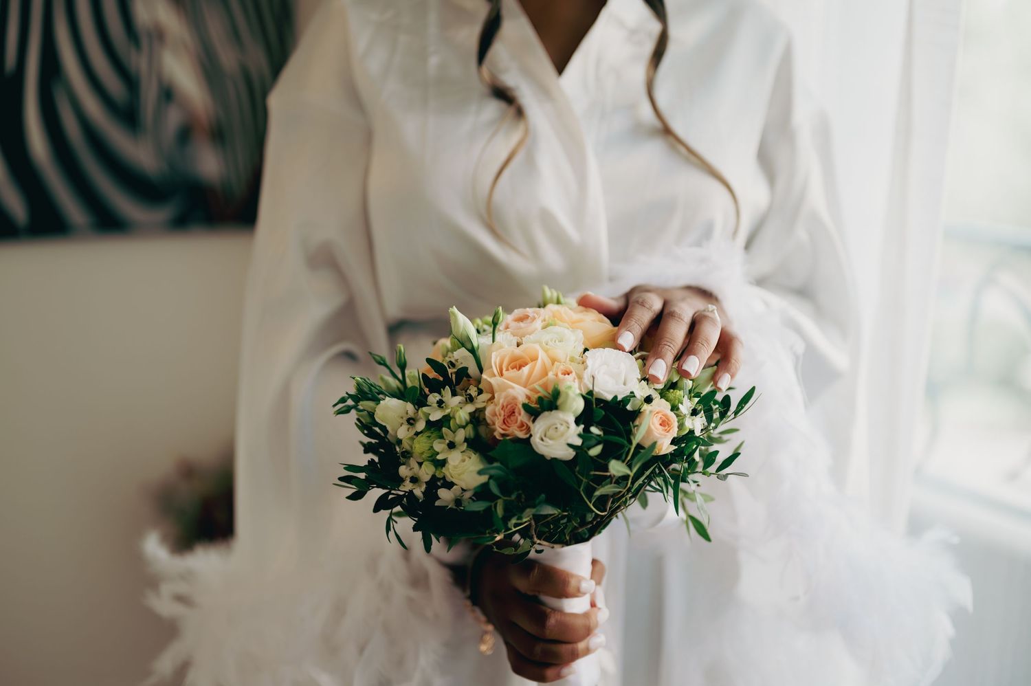 Bride in a white feathered robe holds a delicate bouquet of peach roses, white flowers, and greenery before her wedding.