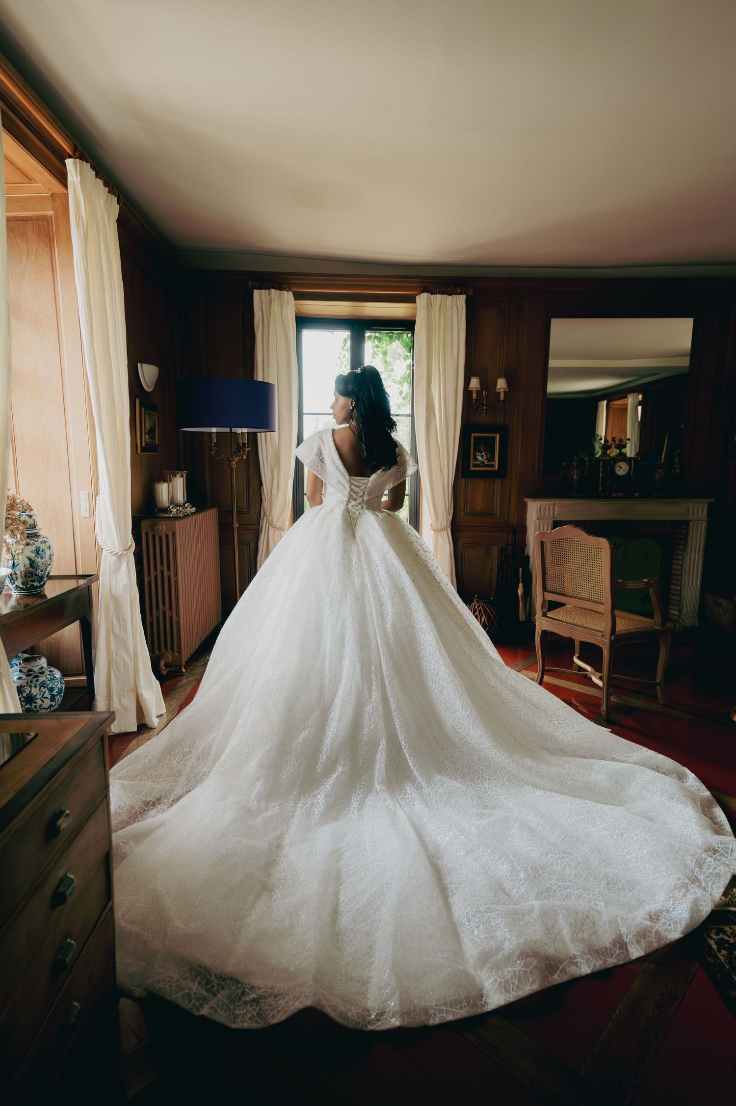 Bride in stunning backless lace ball gown with long cathedral train standing by window in classic wood-paneled room.