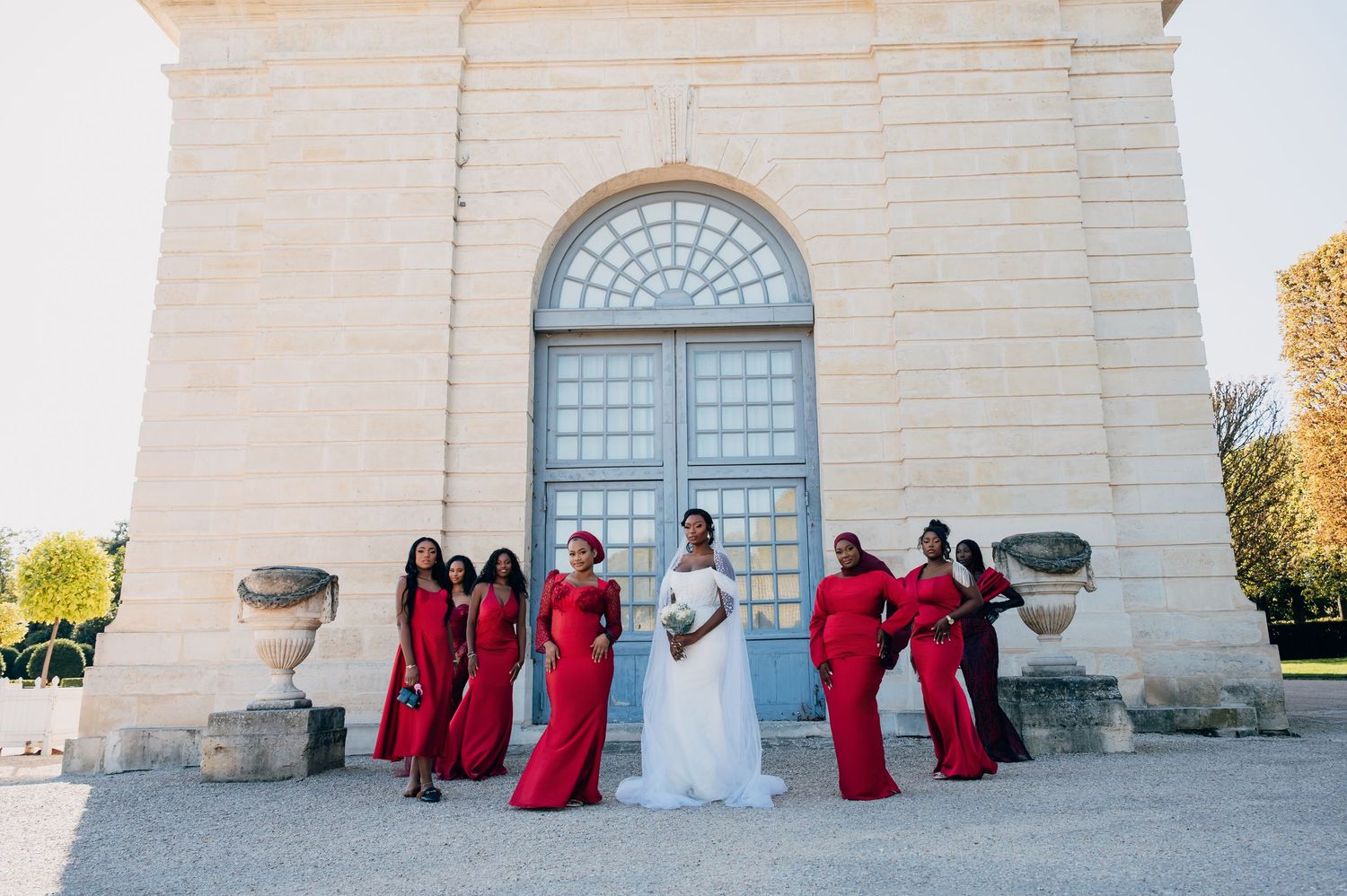 Bride surrounded by bridesmaids in red gowns posing outside a classic stone building.