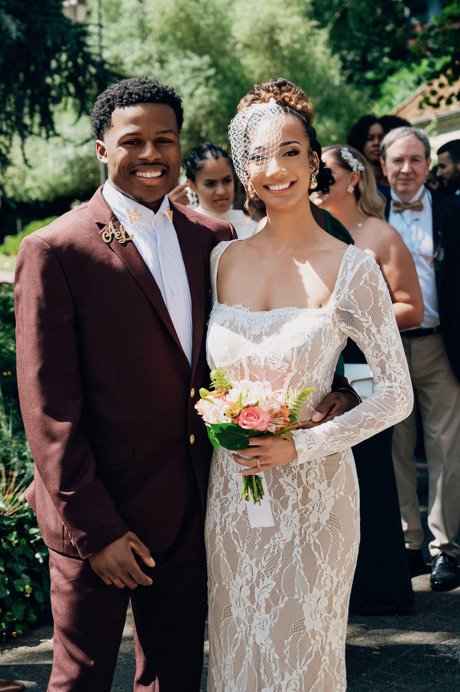 Smiling bride and groom posing together outdoors, she holds pink floral bouquet.