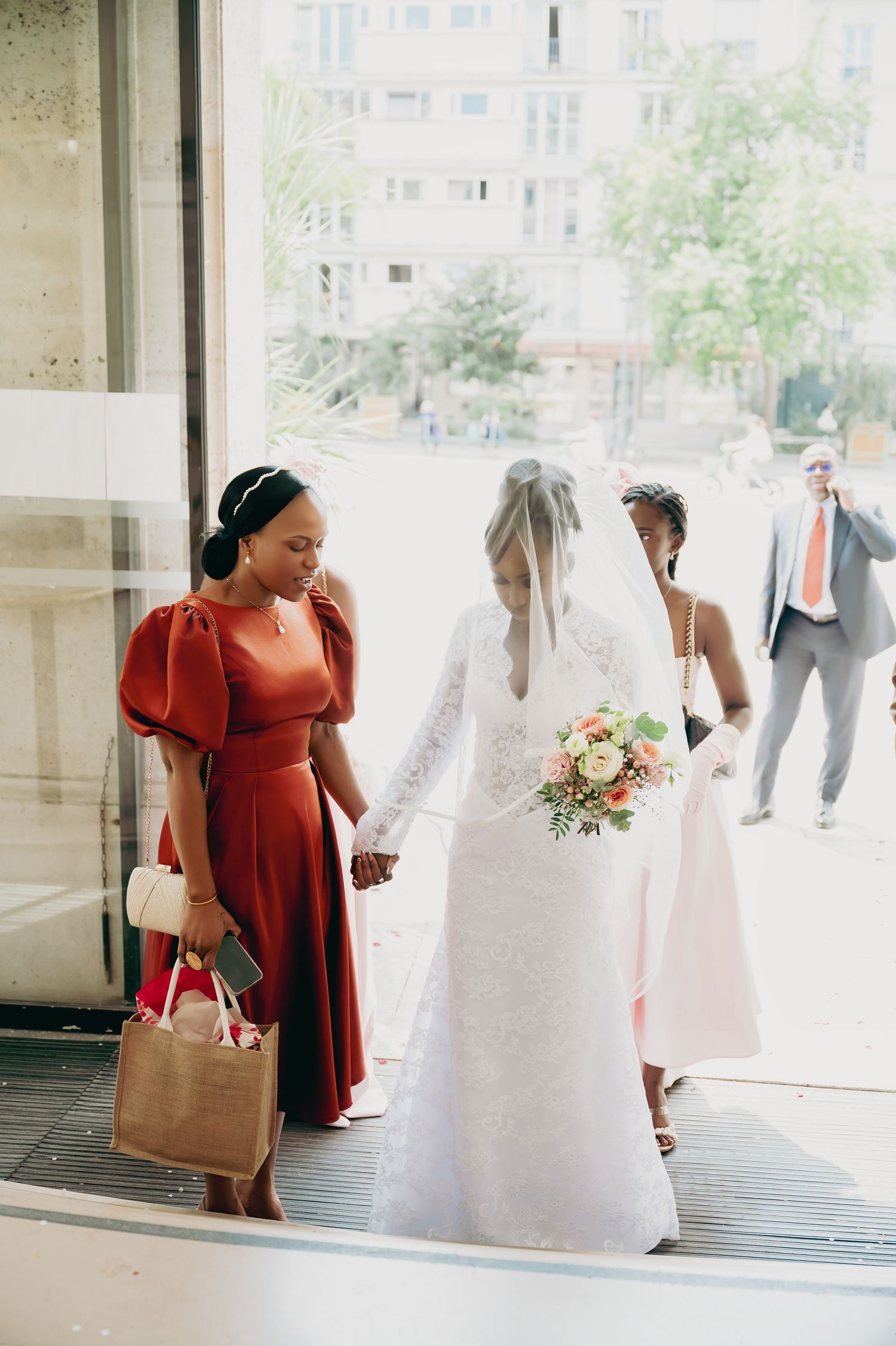 Bride in lace gown and veil walks with bridesmaids in red and pink dresses at wedding.