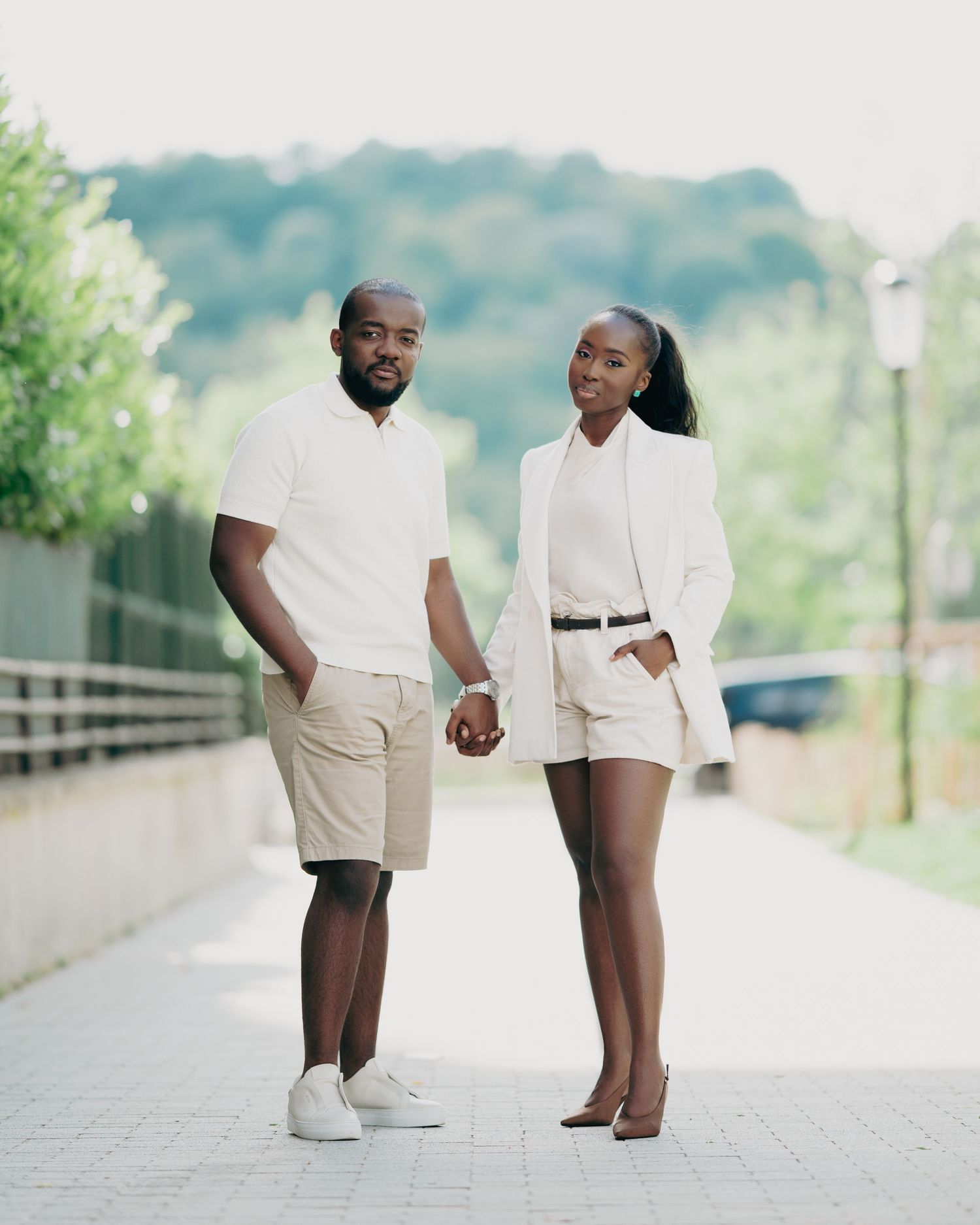 A couple in coordinated white and beige outfits hold hands while posing on a tree-lined walkway.