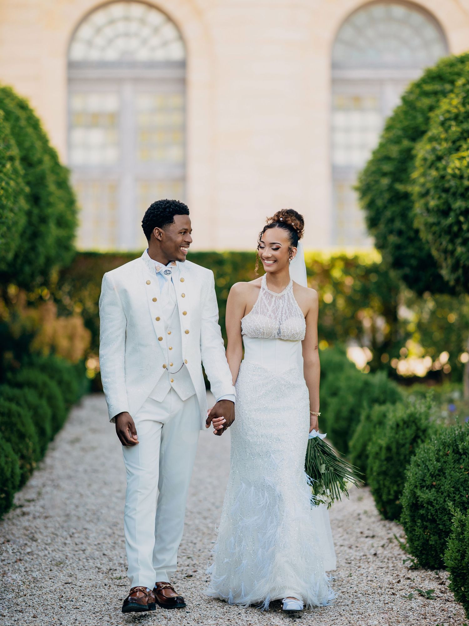 Bride and groom walk hand in hand through elegant garden in matching white wedding attire.