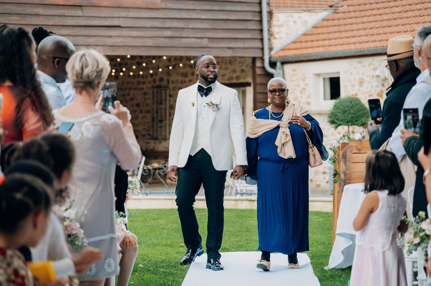 Groom in white tuxedo walking down the aisle with a family member at an outdoor wedding ceremony.