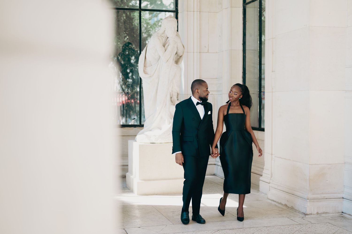 Stylish couple in coordinated dark green formal wear walking hand in hand near a classical white marble statue.