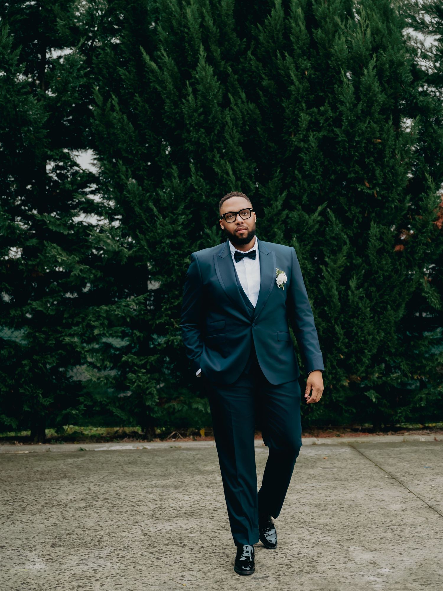 Dapper Black groom in fitted navy tuxedo with bow tie and boutonniere walking confidently before green trees.