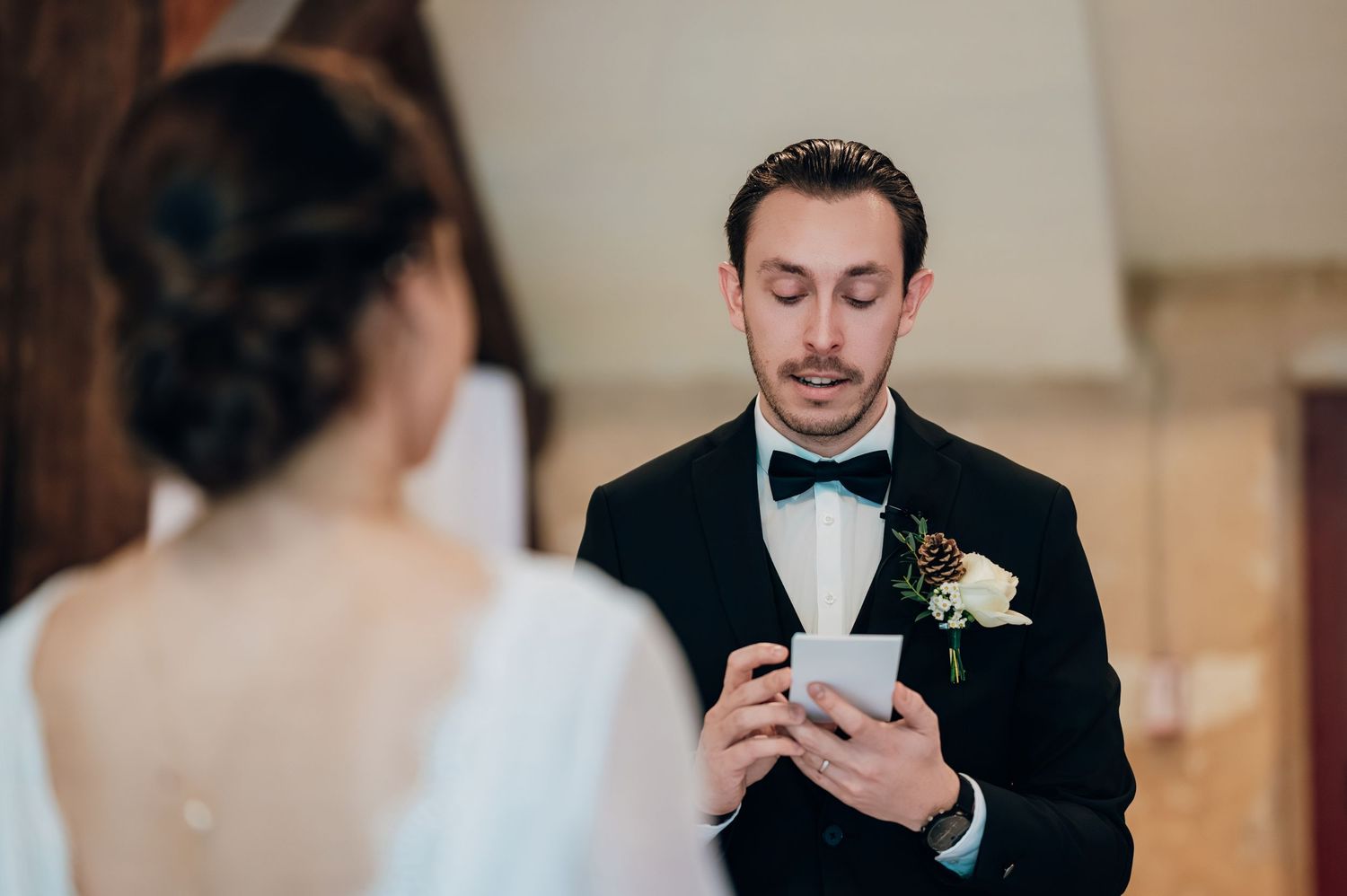 Groom in black tuxedo reading heartfelt wedding vows from card during intimate ceremony.