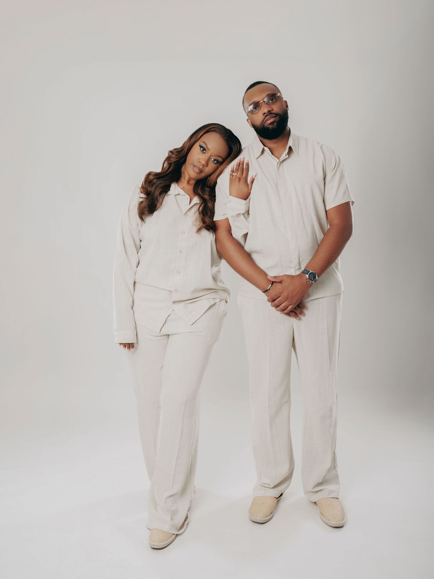 Couple in matching cream outfits pose together in a studio, woman leaning on mans shoulder.