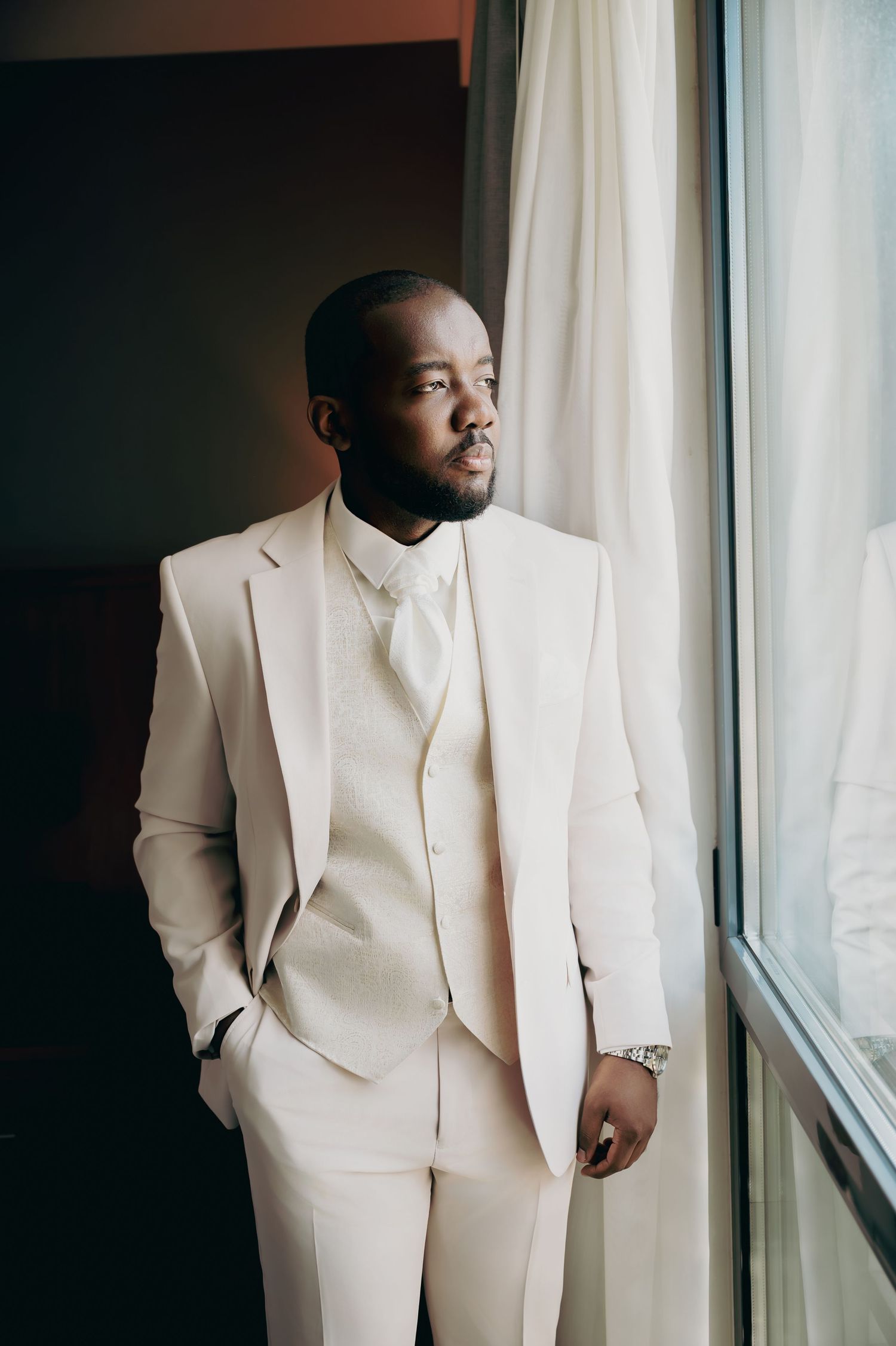 Elegant groom in cream three-piece suit standing by window in sophisticated wedding portrait.
