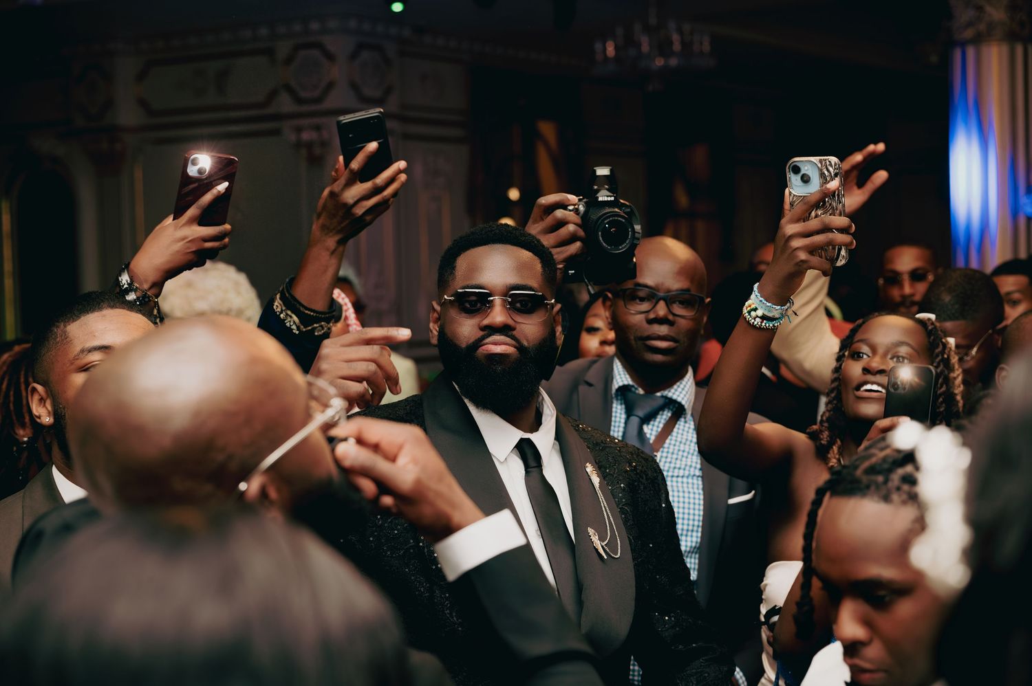 Wedding guests in formal attire celebrate and take photos with phones at a lively elegant wedding reception.