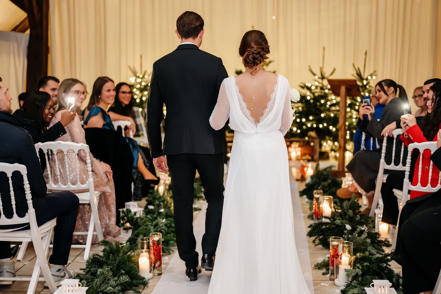 Bride and groom walking down the aisle lined with candles and greenery, guests applauding in a warmly lit venue.