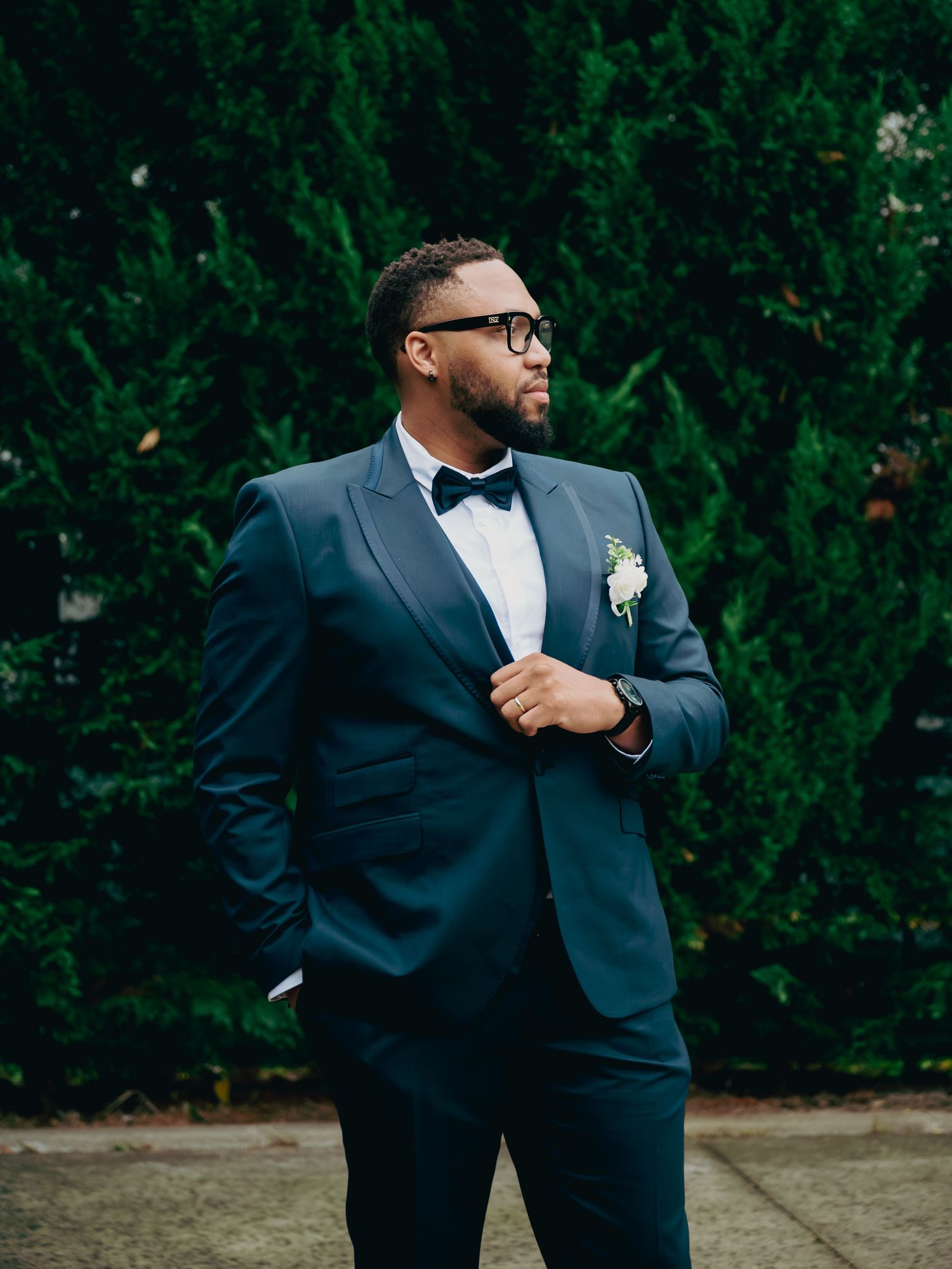Handsome groom in navy tuxedo with bow tie and boutonniere posing confidently in front of dark hedges.