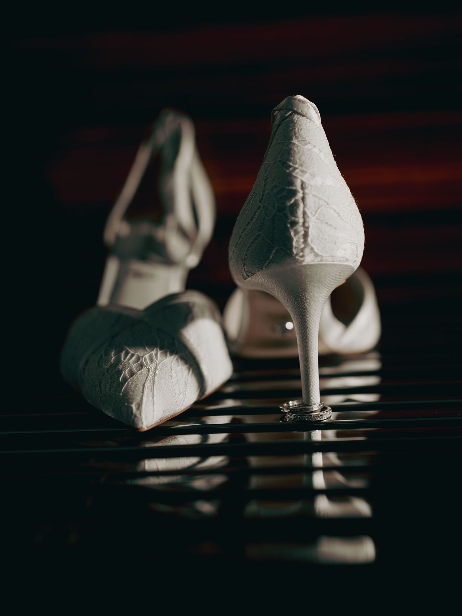 White bridal heels with diamond wedding rings resting beneath them in dramatic lighting.