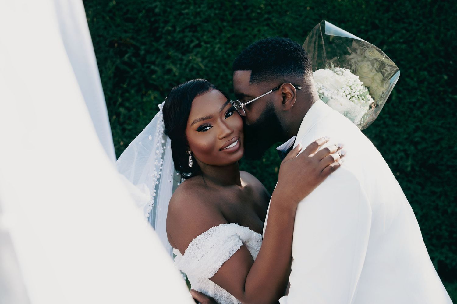 Groom tenderly kisses brides cheek as she smiles radiantly holding white bouquet under veil.