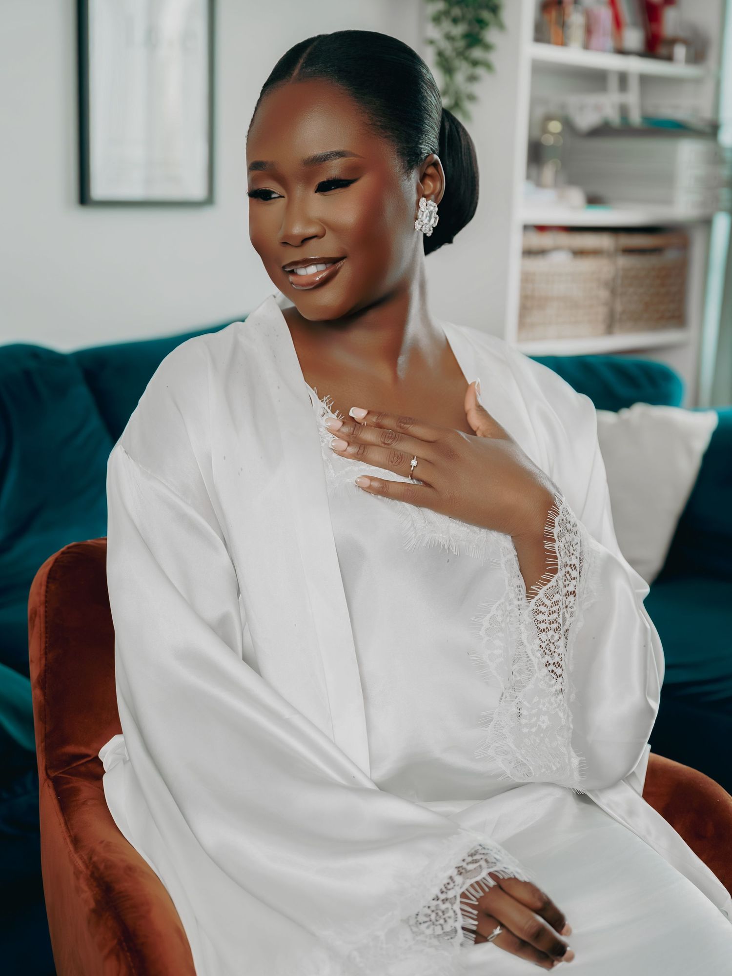 Radiant bride in white lace robe sits in a velvet chair, smiling and touching her necklace during prep.