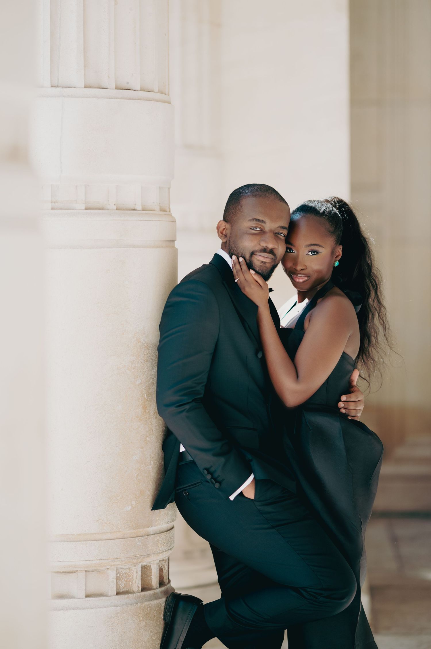 Elegant Black couple in matching black formal wear embrace beside a stone column.