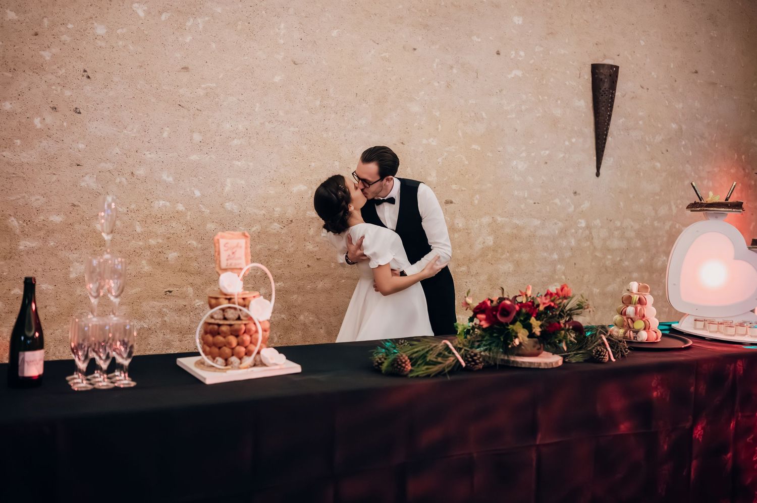 Bride and groom sharing a kiss behind wedding dessert table with champagne and floral arrangements.