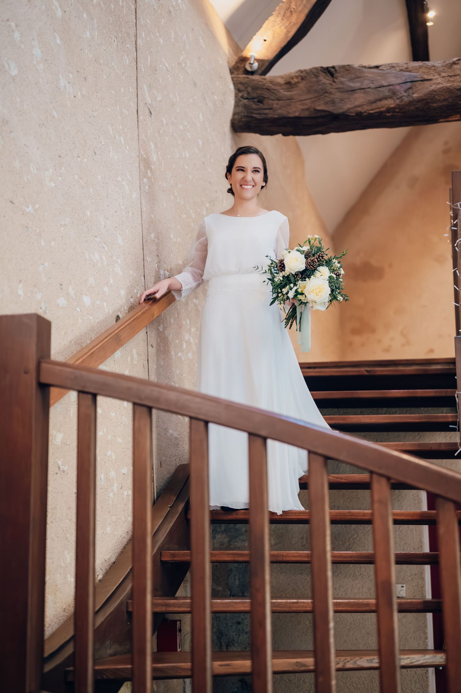 Beautiful bride in long-sleeve white wedding gown holding a white floral bouquet while posing on wooden staircase.