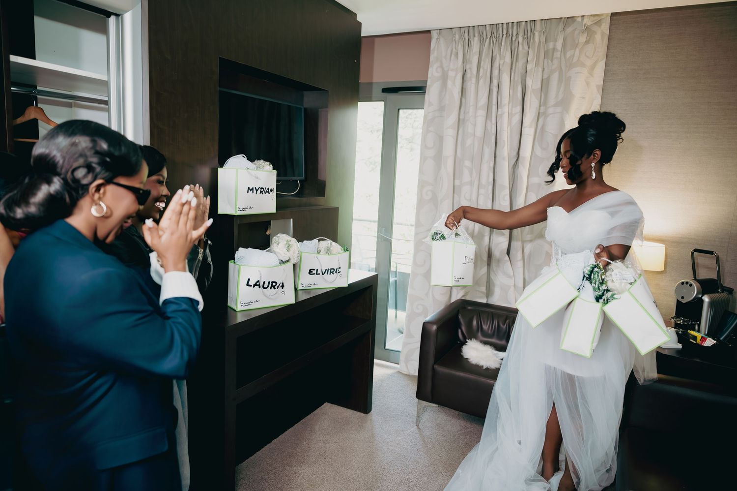 Bride in white gown surprising bridesmaids with personalized gift bags in a hotel room.