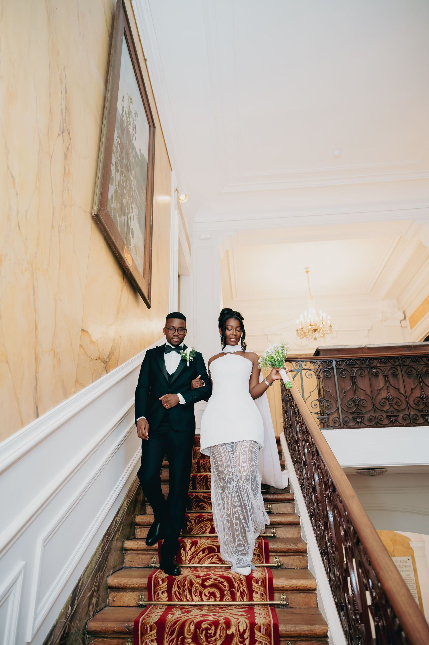 Newlywed couple walks hand in hand down grand red-carpeted staircase in elegant French chateau venue.