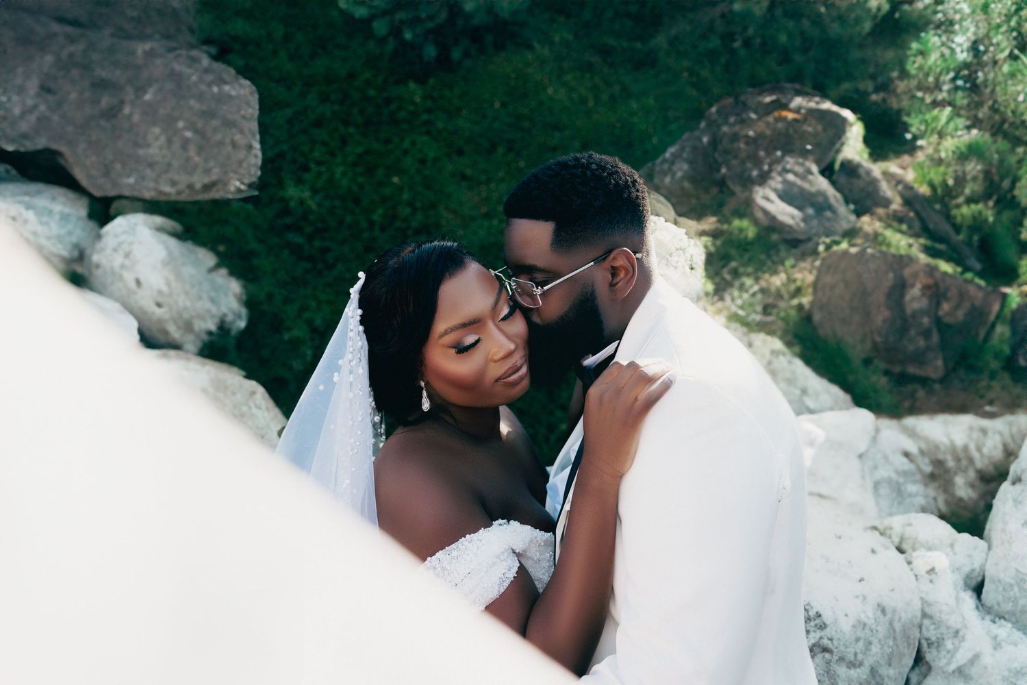 Intimate bride and groom portrait among rocks, bride in off-shoulder gown with flowing veil.