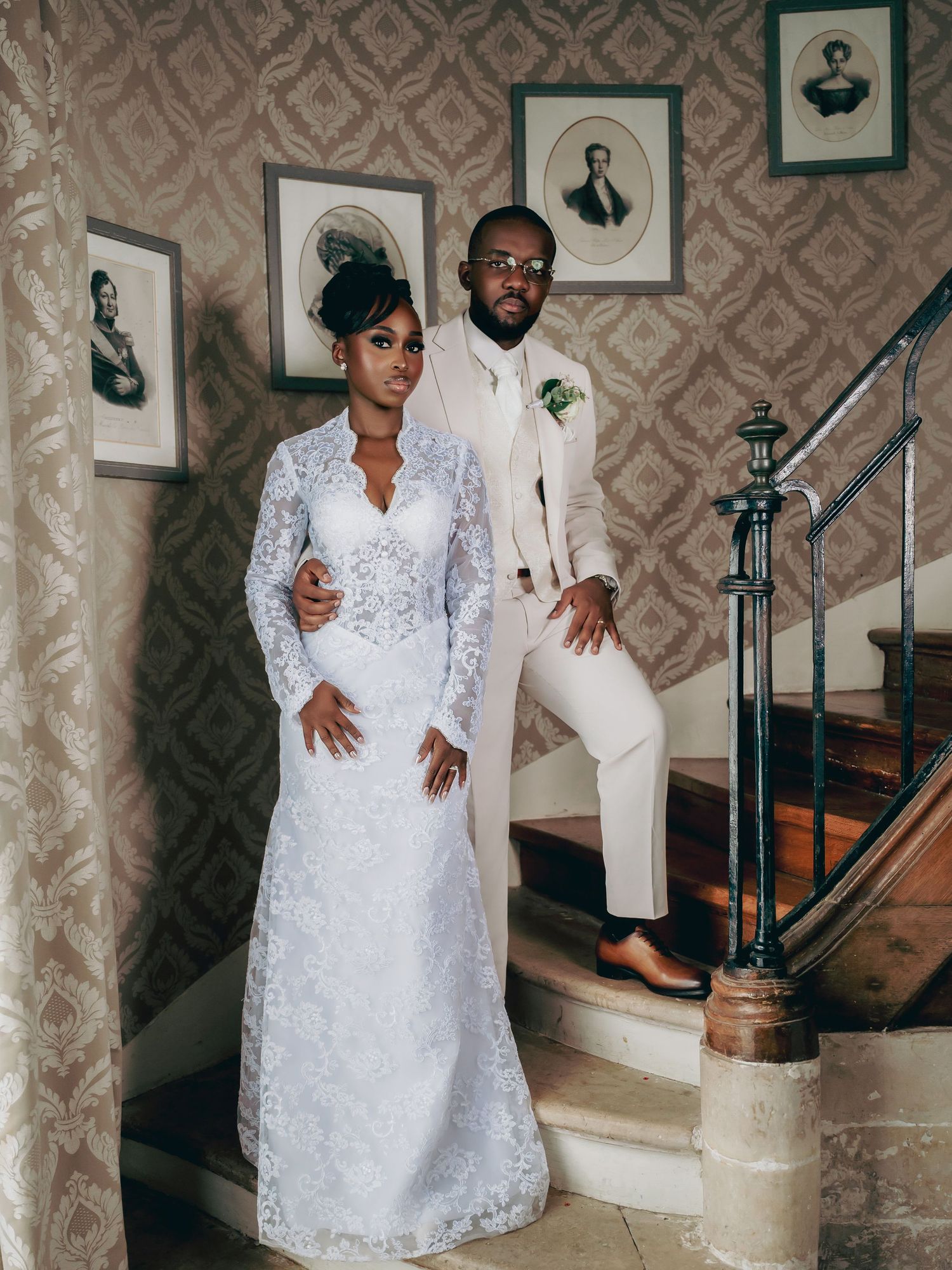 Bride in long-sleeve lace wedding gown and groom in cream suit pose on classic interior staircase.