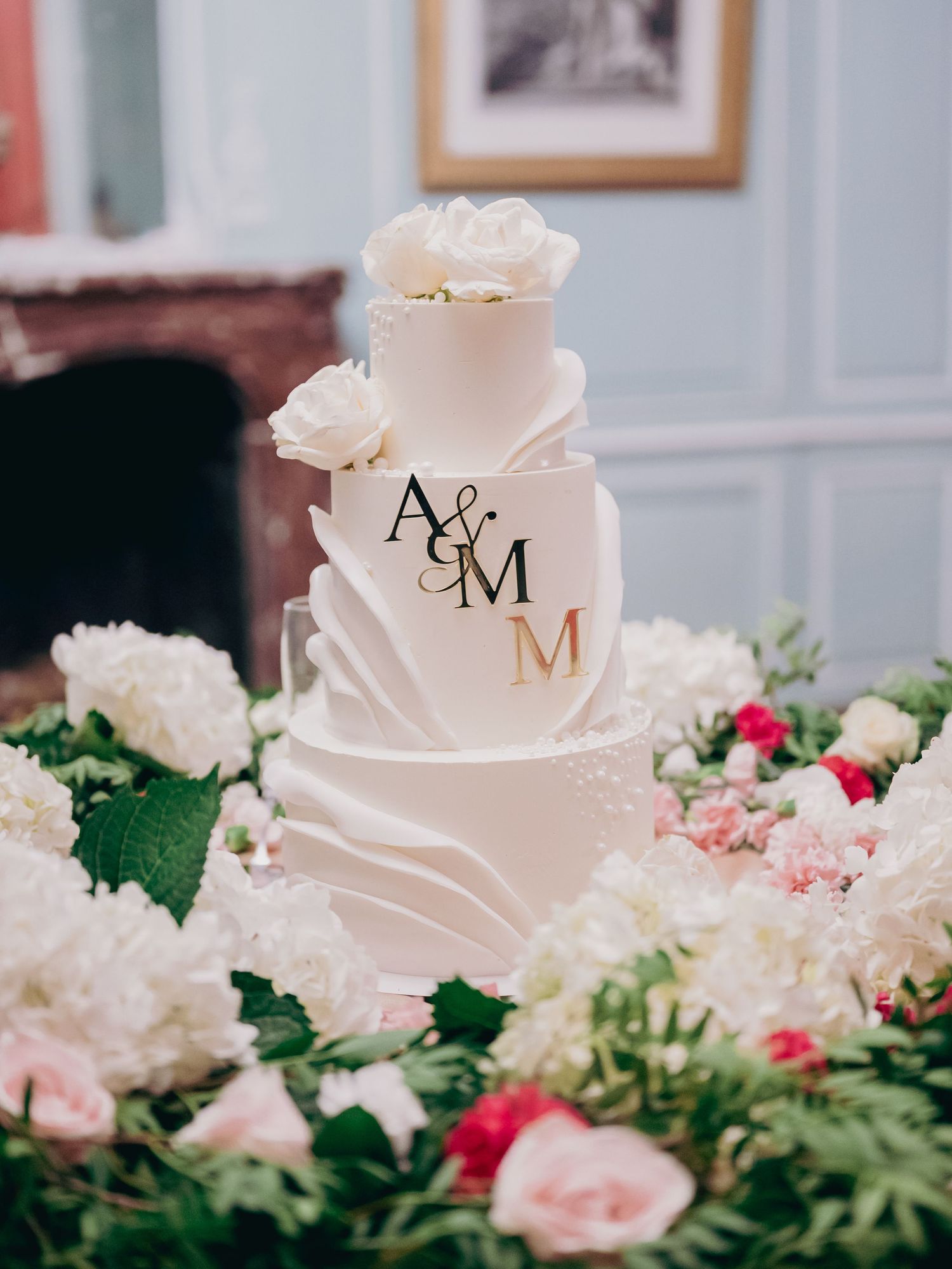 Elegant three-tier white wedding cake with gold A&M monogram surrounded by white and pink flowers.