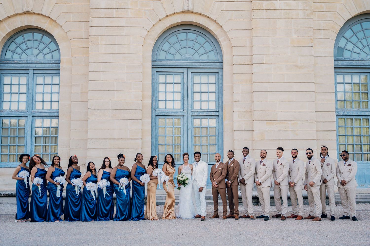 Wedding party lineup featuring bridesmaids in blue dresses and groomsmen in tan suits before a classical stone building.