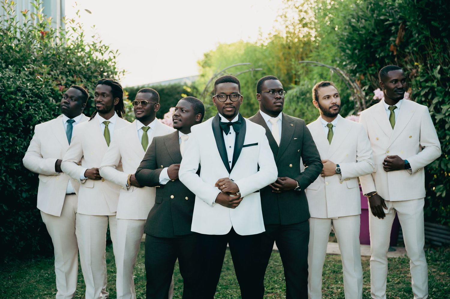 Groom in white tuxedo stands with groomsmen in green and cream suits outdoors.