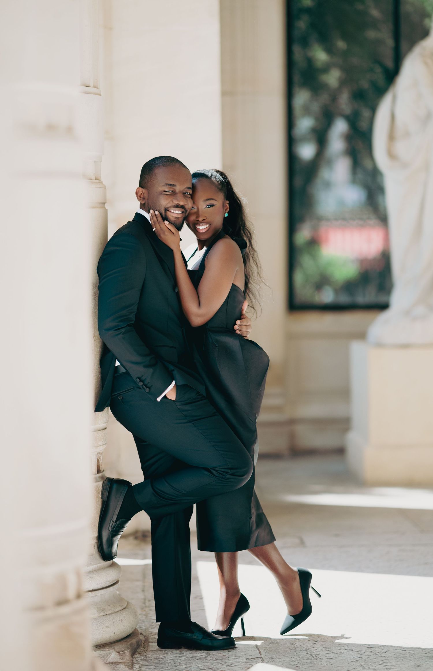 Joyful Black couple in black formal wear laughing and embracing between elegant stone columns outdoors.