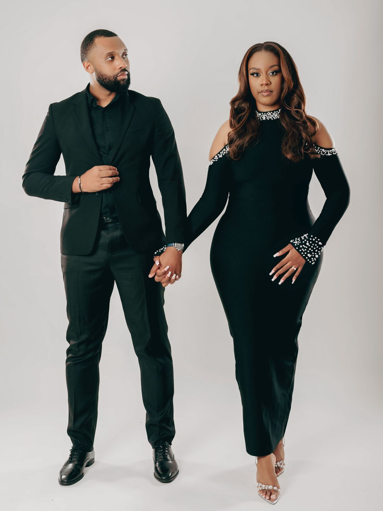 Elegant couple in coordinating black outfits holding hands in a full-length studio portrait photograph.