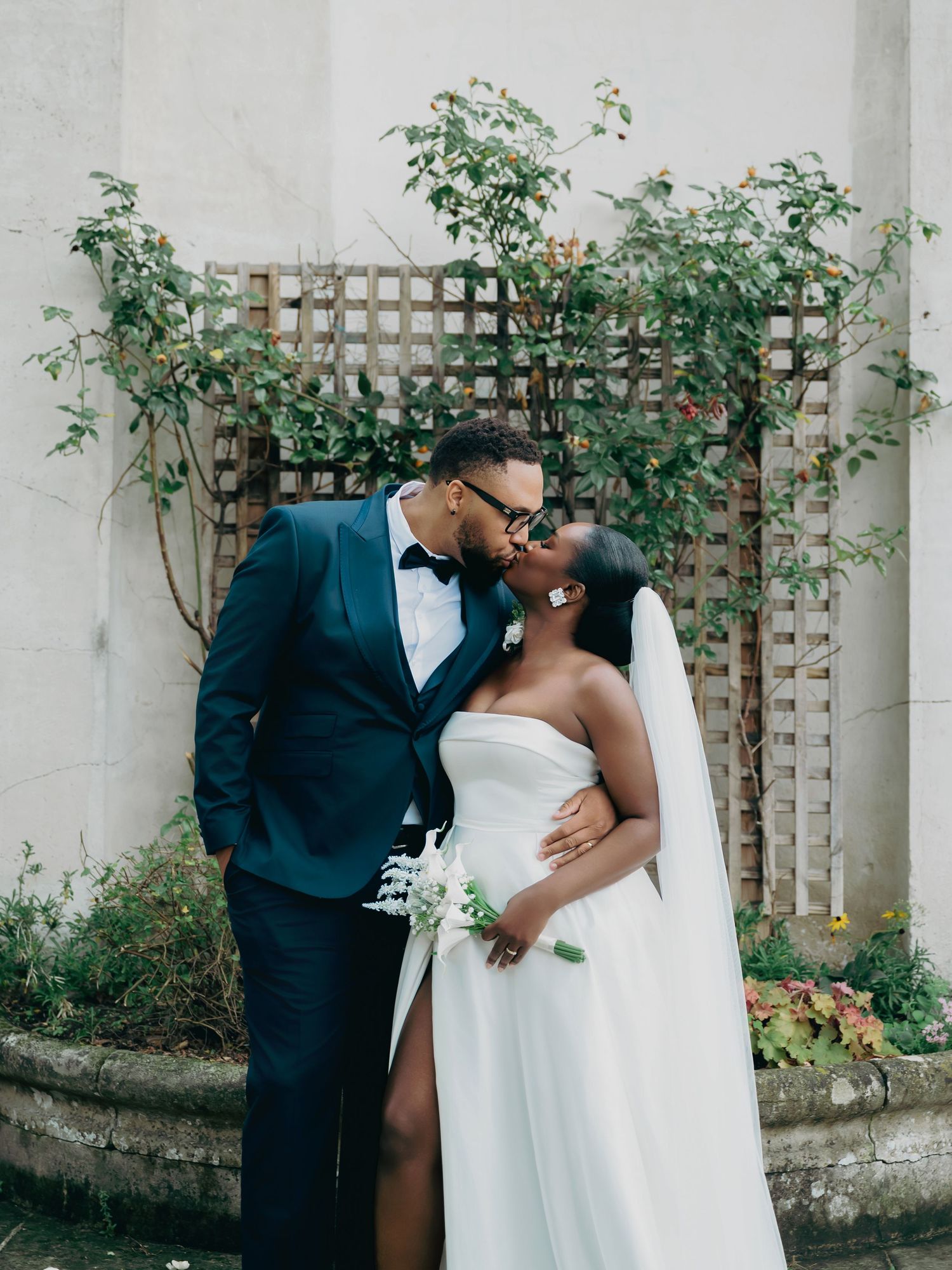 Newlyweds kissing tenderly in front of lattice wall with greenery in outdoor wedding portrait.