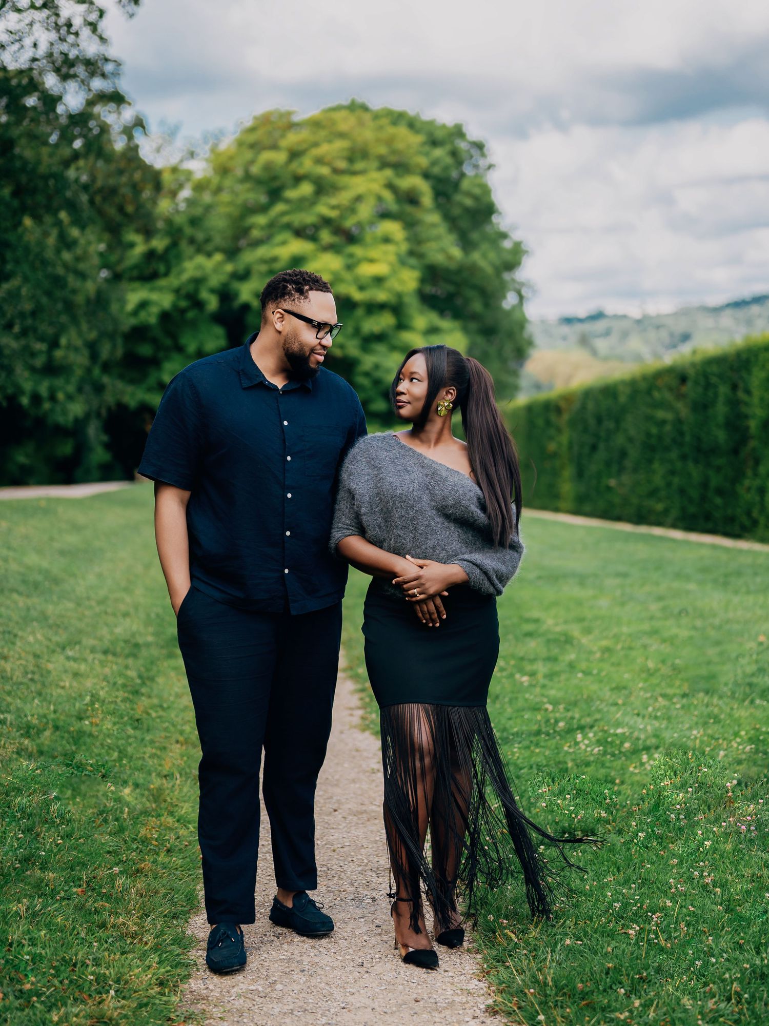 Couple walking together on garden path, smiling at each other, woman wearing gray wrap and black fringe skirt.
