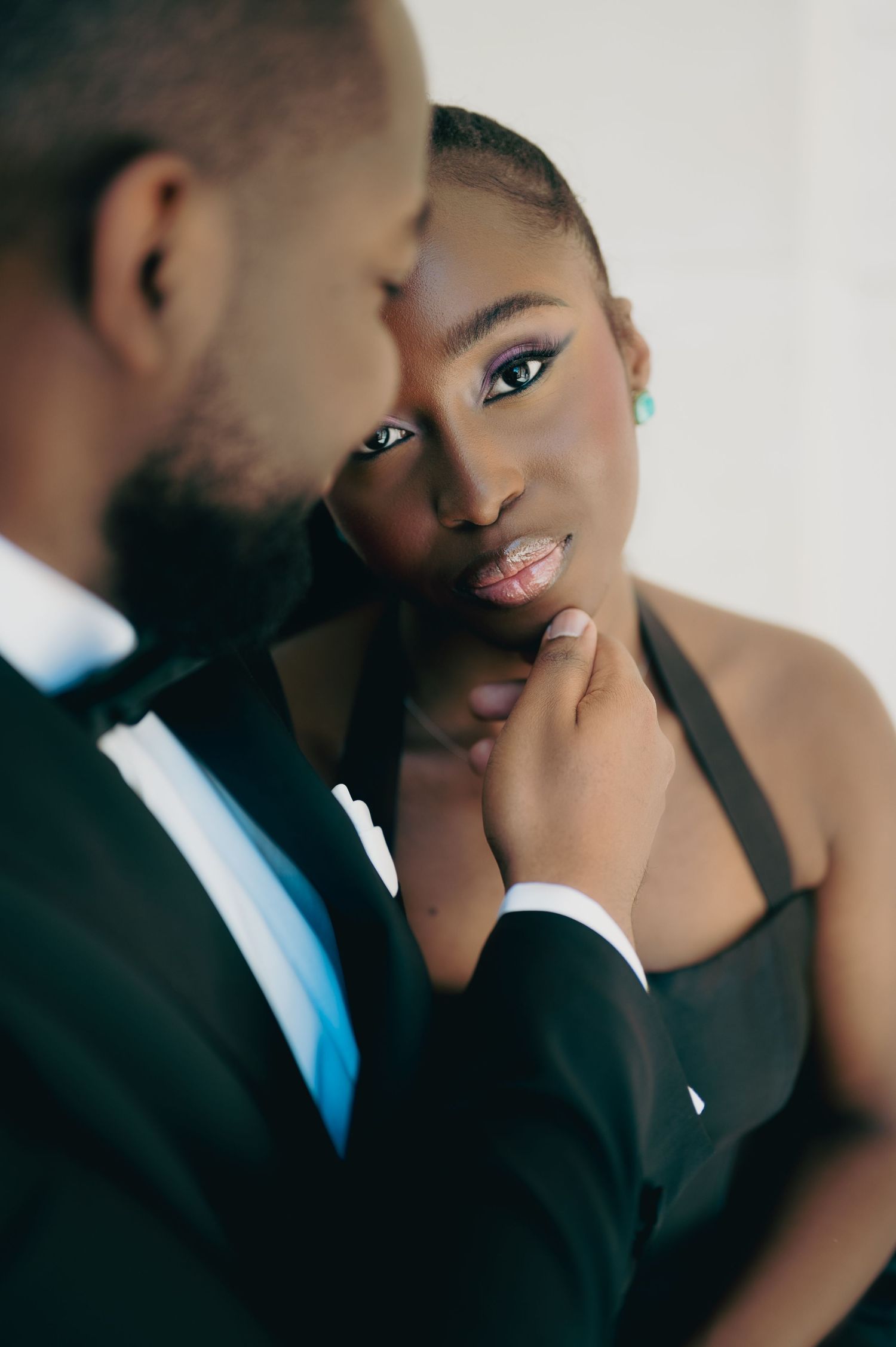 Black woman in black halter dress gazes intensely as man in tuxedo gently holds her chin.