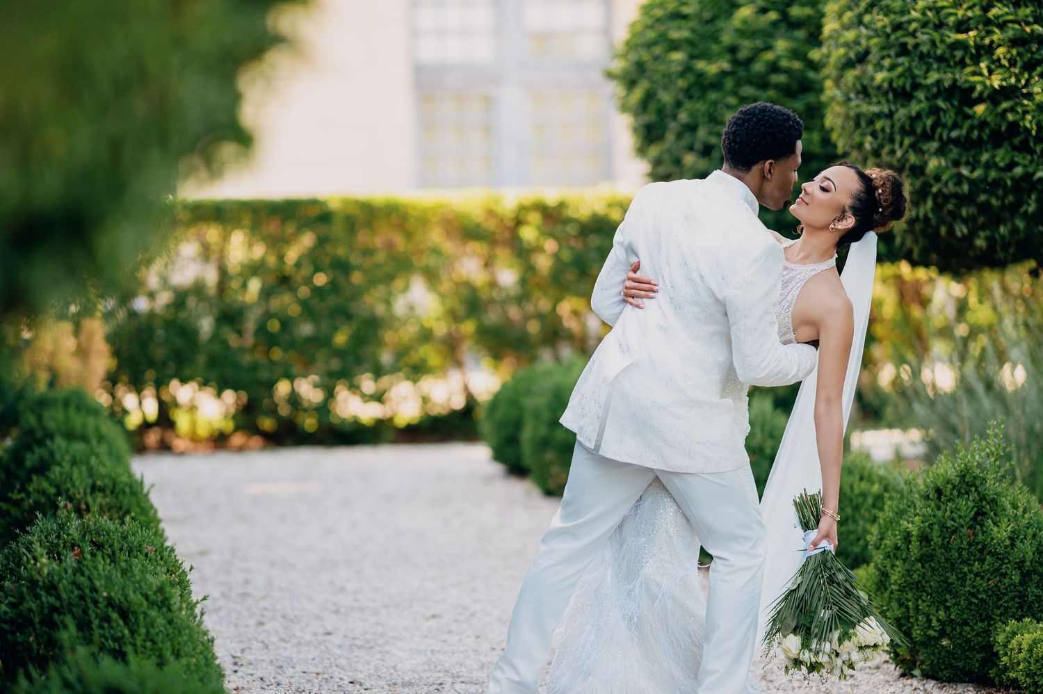 Interracial couple sharing a romantic kiss in a lush garden, groom in white suit and bride in elegant wedding gown.