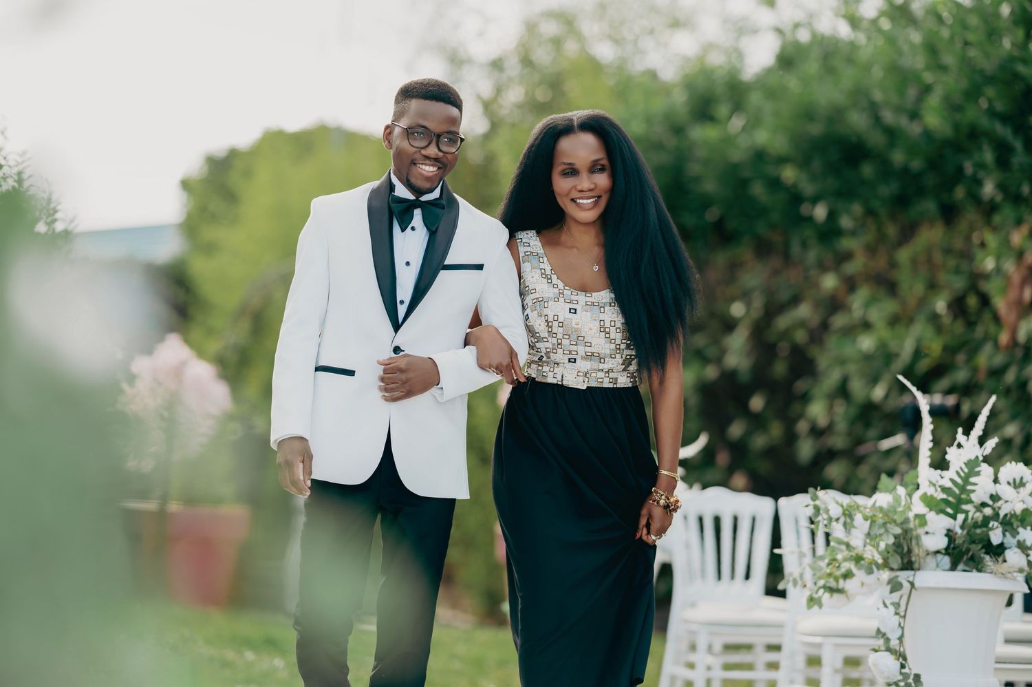 A smiling couple poses together outdoors, man in white tuxedo and woman in elegant black and gold outfit.
