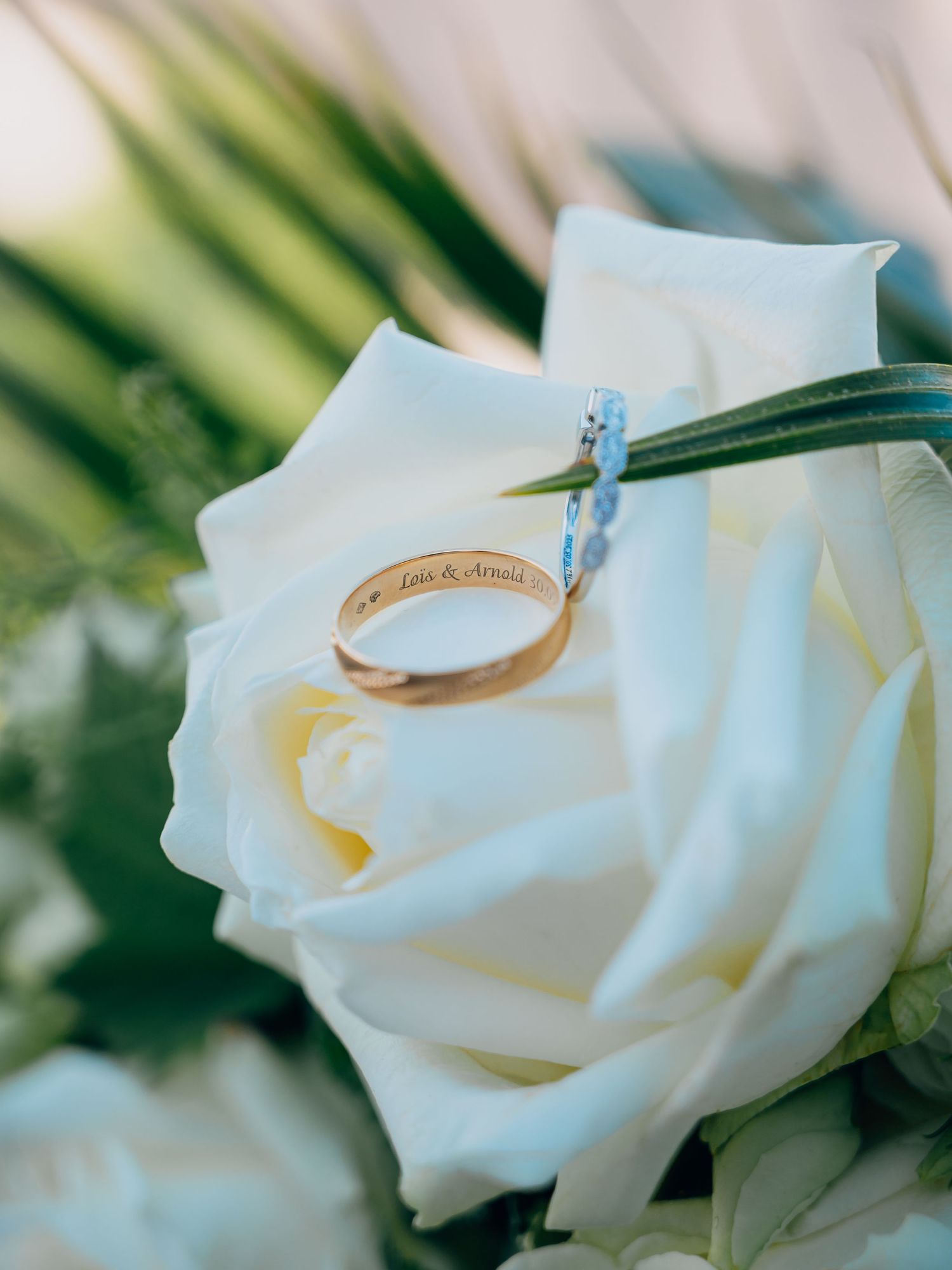 Gold engraved wedding band resting on a white rose with a diamond ring, personalized with couple's names.